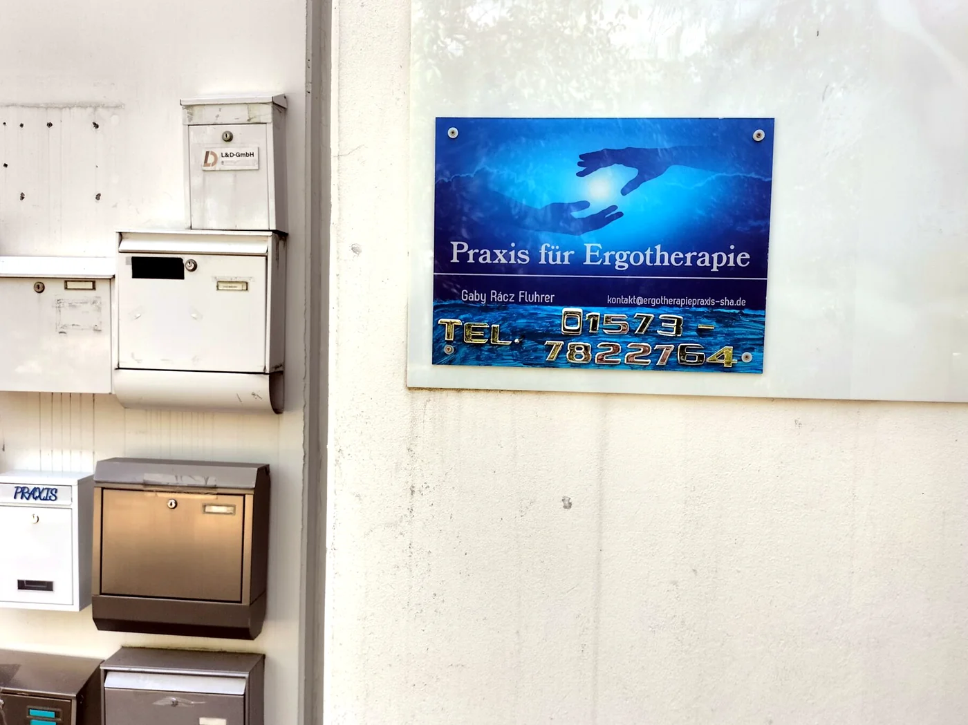 Blaues Schild „Praxis für Ergotherapie Gaby Rácz-Fluhrer“ mit Telefonnummer neben den Briefkästen am Eingang